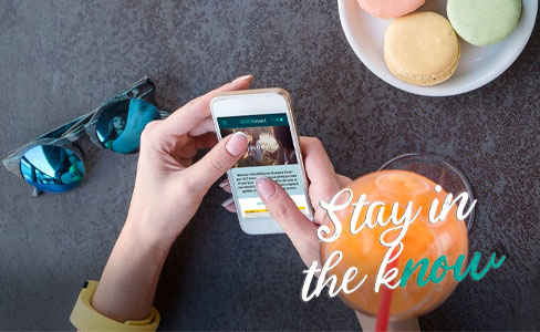 A person uses a smartphone at a table with sunglasses, colorful macarons on a plate, and an orange beverage. The text “Stay in the know” appears in stylish white and blue lettering.