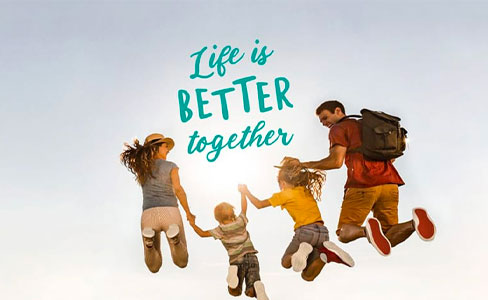 Four people—two adults and two children—are holding hands and jumping mid-air against a clear sky. The text above them reads, “Life is BETTER together.”.