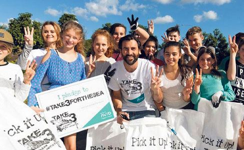 A group of smiling people, including children and adults, hold bags and make peace signs while participating in a litter clean-up event under a blue sky. The bags read "Pick it Up" and shirts display "TAKE 3 FOR THE SEA".