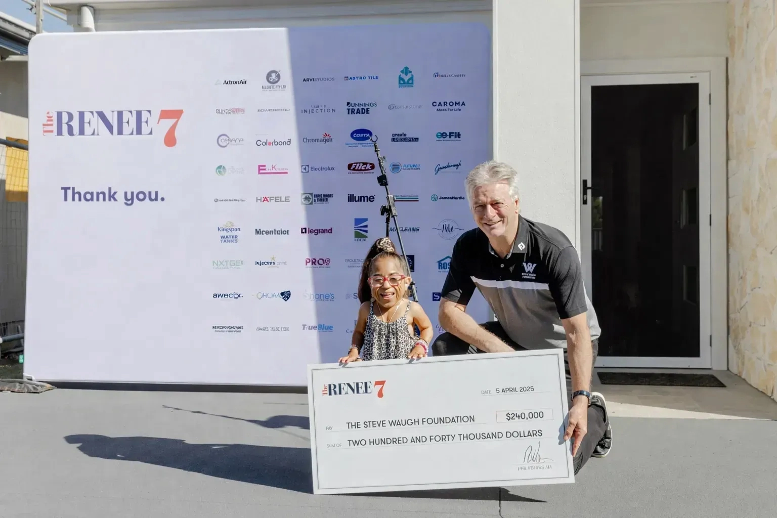 A smiling girl and a man kneel outside, holding a large donation check for $240,000 to The Steve Waugh Foundation. Behind them is a sponsor banner with logos and "Thank you" written on it.
