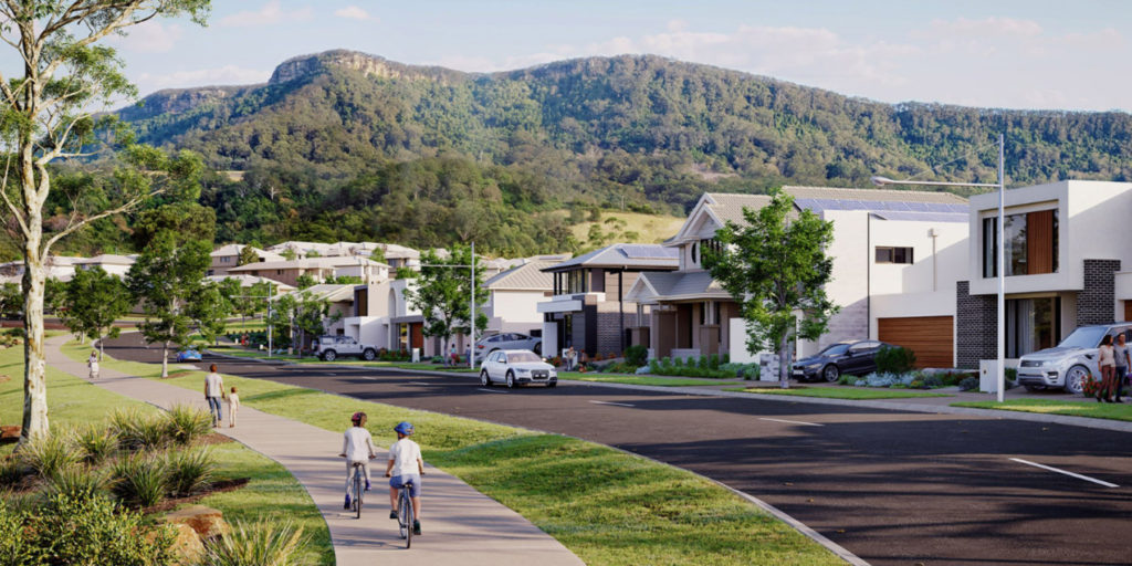A suburban street with modern houses, trees, and parked cars, set against a backdrop of green mountains. Two children ride bikes on a sidewalk while an adult walks nearby under a sunny, clear sky.