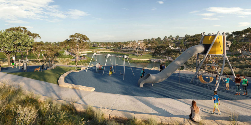 A modern playground with swings, a large slide, and climbing structures. Children play on the equipment while adults watch nearby. The playground is surrounded by grass, trees, and open parkland under a blue sky.