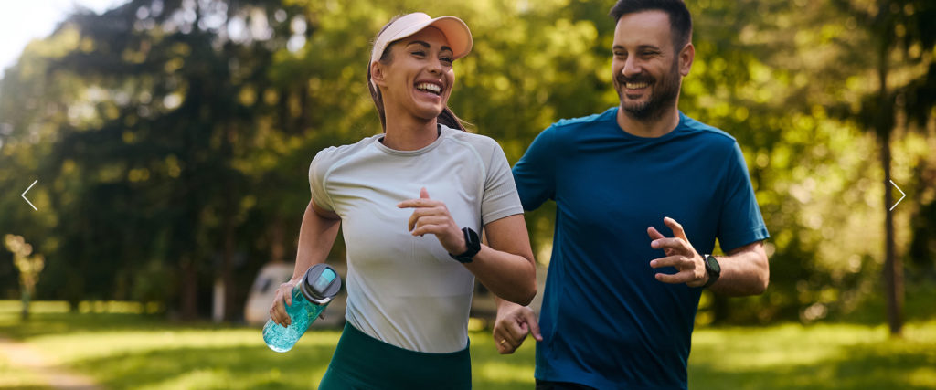 A woman and a man smiling and jogging together in a sunny park, surrounded by green trees. The woman holds a water bottle and wears a cap, while both wear athletic clothing.