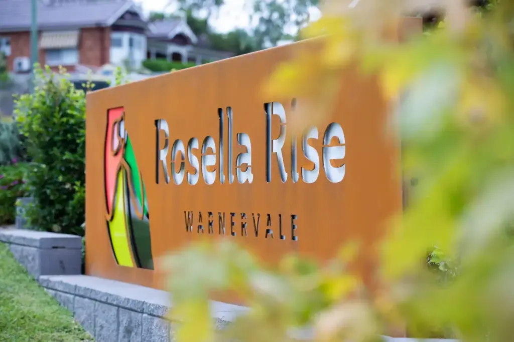 A large orange sign with the words "Rosella Rise Warnervale" and a colorful parrot graphic, surrounded by green foliage and a blurred house in the background.